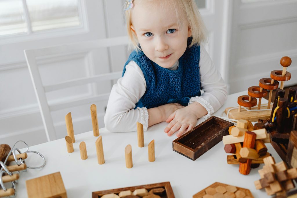 pexels-photo-7296786-7296786 A Young Girl Smiling while Sitting Near the Table with Wooden Toys